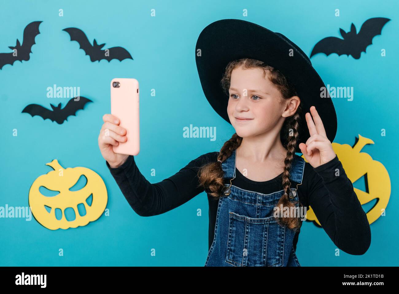 Beautiful little girl kid in black hat holding telephone shooting making funky selfies showing v-sign symbol, isolated on blue color background in stu Stock Photo