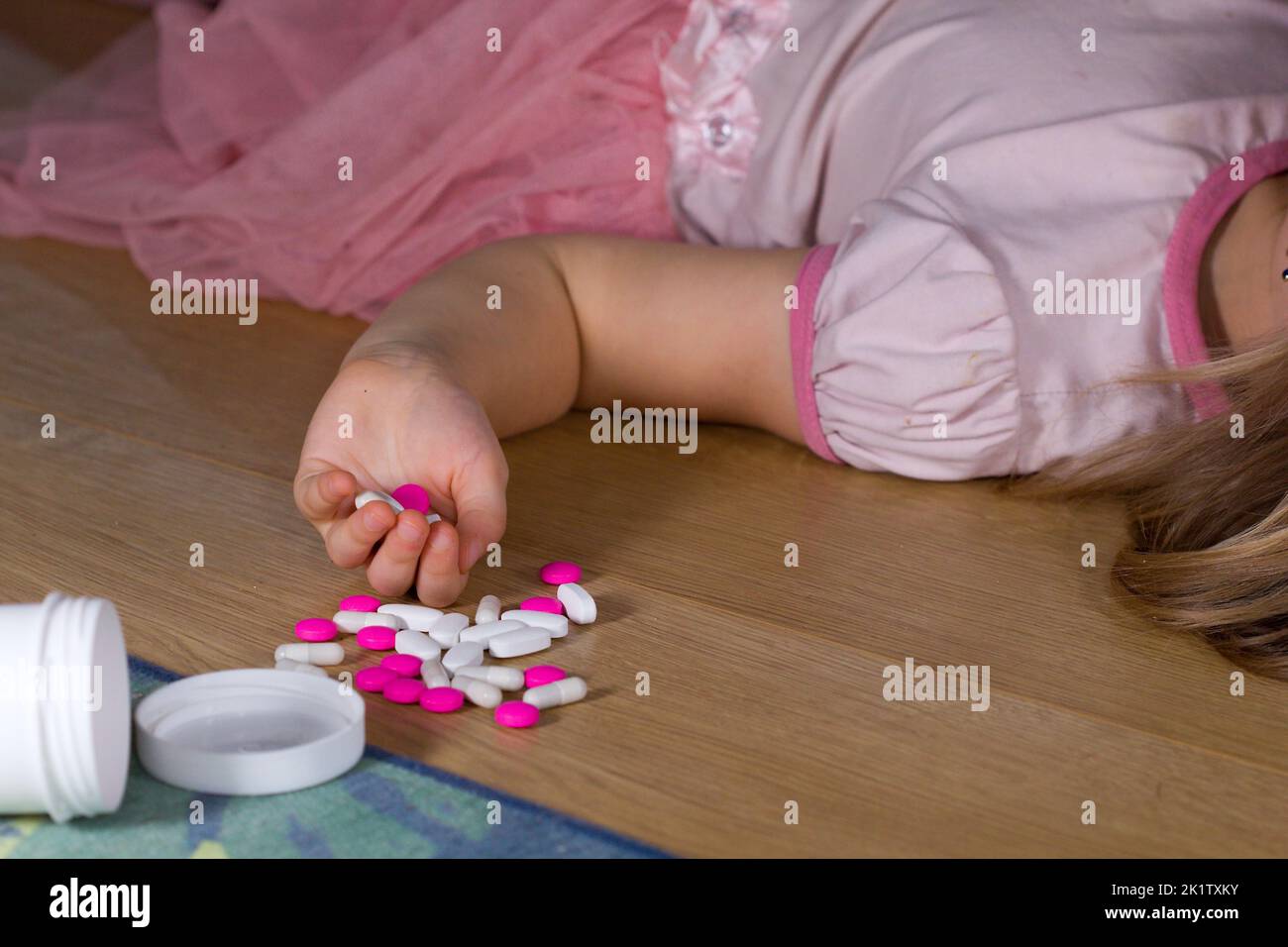 Unconscious little child (girl) with pink pills laying on the floor at home. Danger of medicament intoxication. Stock Photo