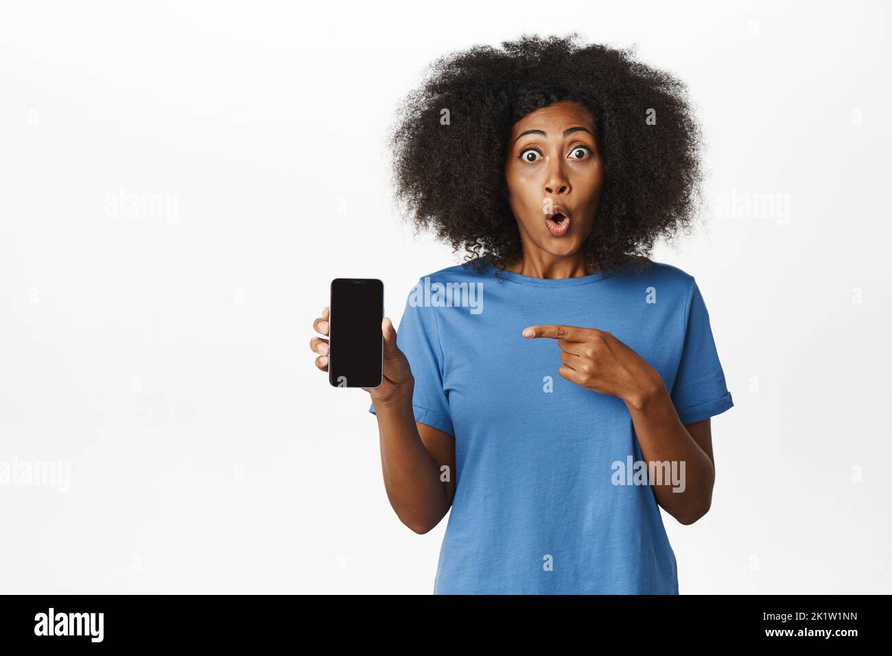 Portrait of amazed and surprised Black woman pointing finger at mobile phone screen, showing smartphone application with enthusiastic face, white back Stock Photo