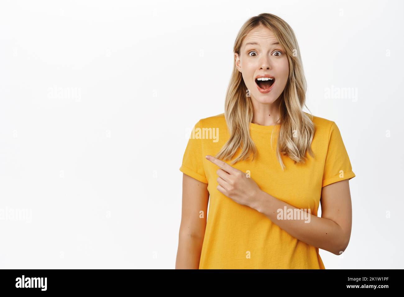 Smiling beautiful blond girl showing announcement, pointing at advertisement, empty copy space, standing in yellow t-shirt over white background. Stock Photo