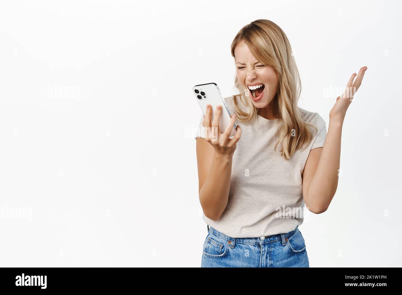 Excited woman looking at mobile phone screen and shouting, screaming at smartphone display, standing over white background. Stock Photo