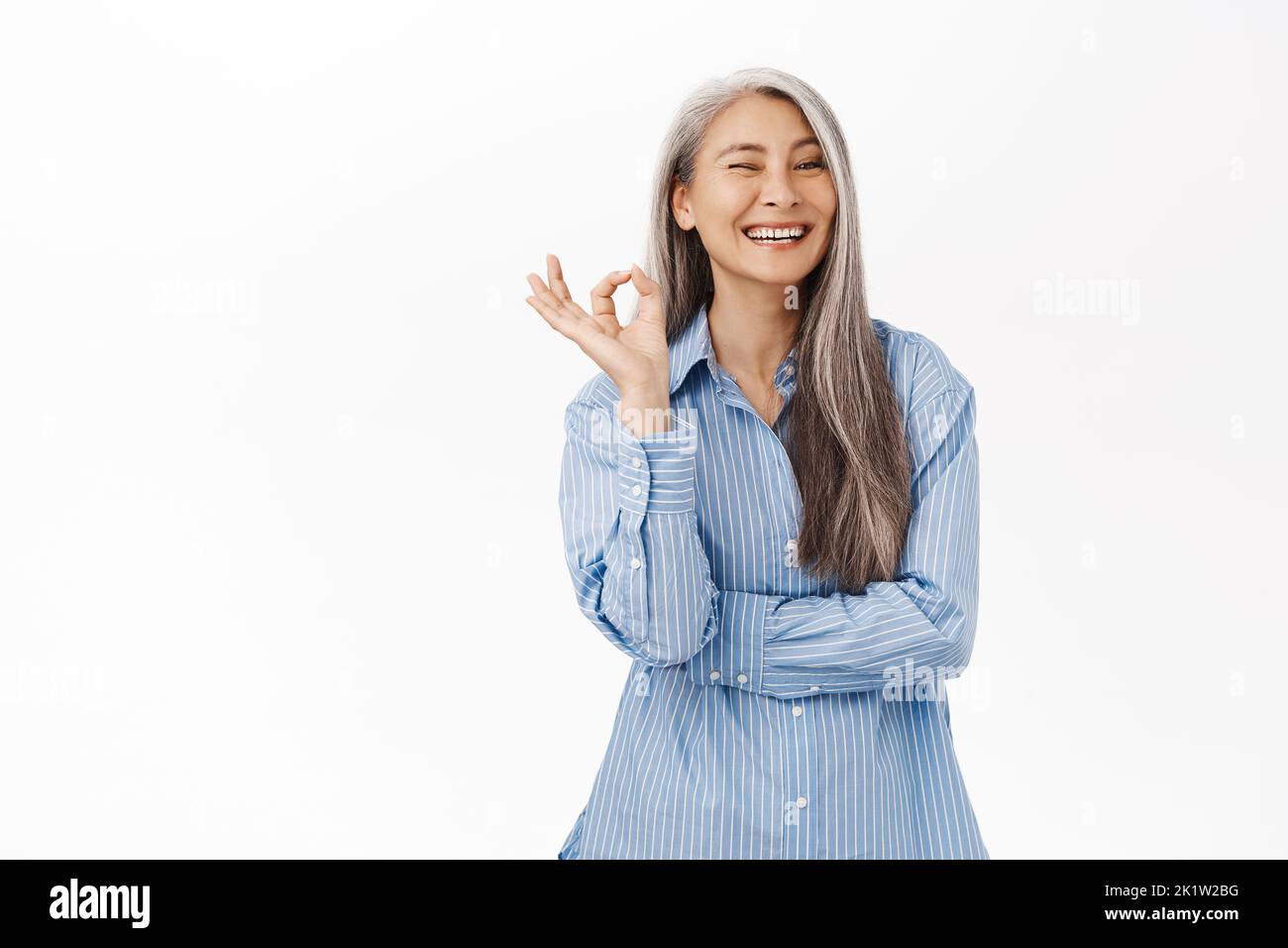 Portrait of beautiful, healthy senior japanese woman, old asian lady smiling, rate smth excellent, make okay, ok sign, standing over white background. Stock Photo