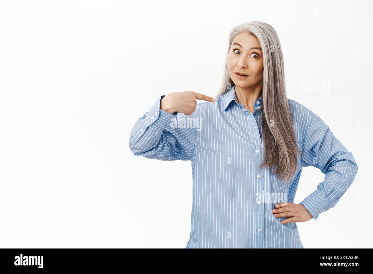 Surprised old asian lady, mother with gray hair, pointing at herself with disbelief, standing over white studio background Stock Photo
