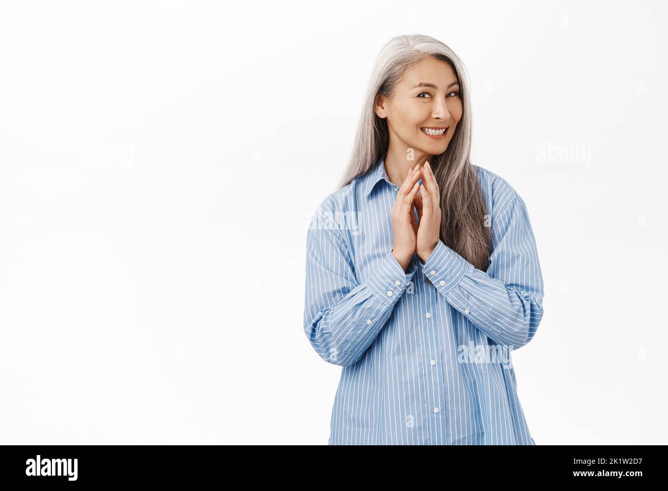 Smiling old asian lady, senior woman scheming, has plan, thinking of something with cunning, coy smile, steeple fingers, standing over white backgroun Stock Photo