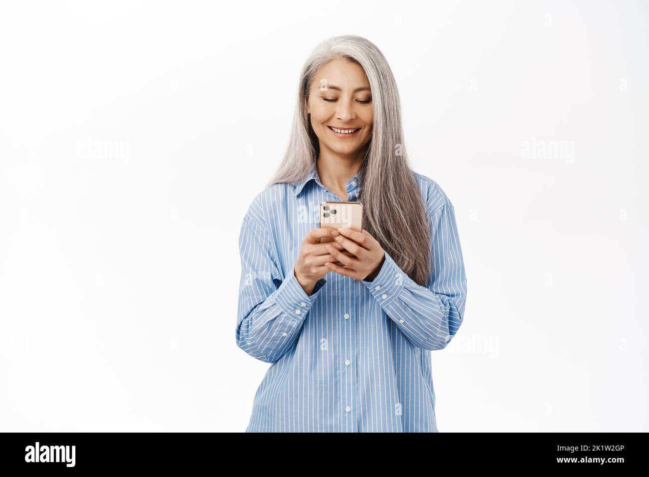 Portrait of smiling senior asian woman using smartphone. Japanese old lady holding mobile phone with happy face, standing over white background. Stock Photo