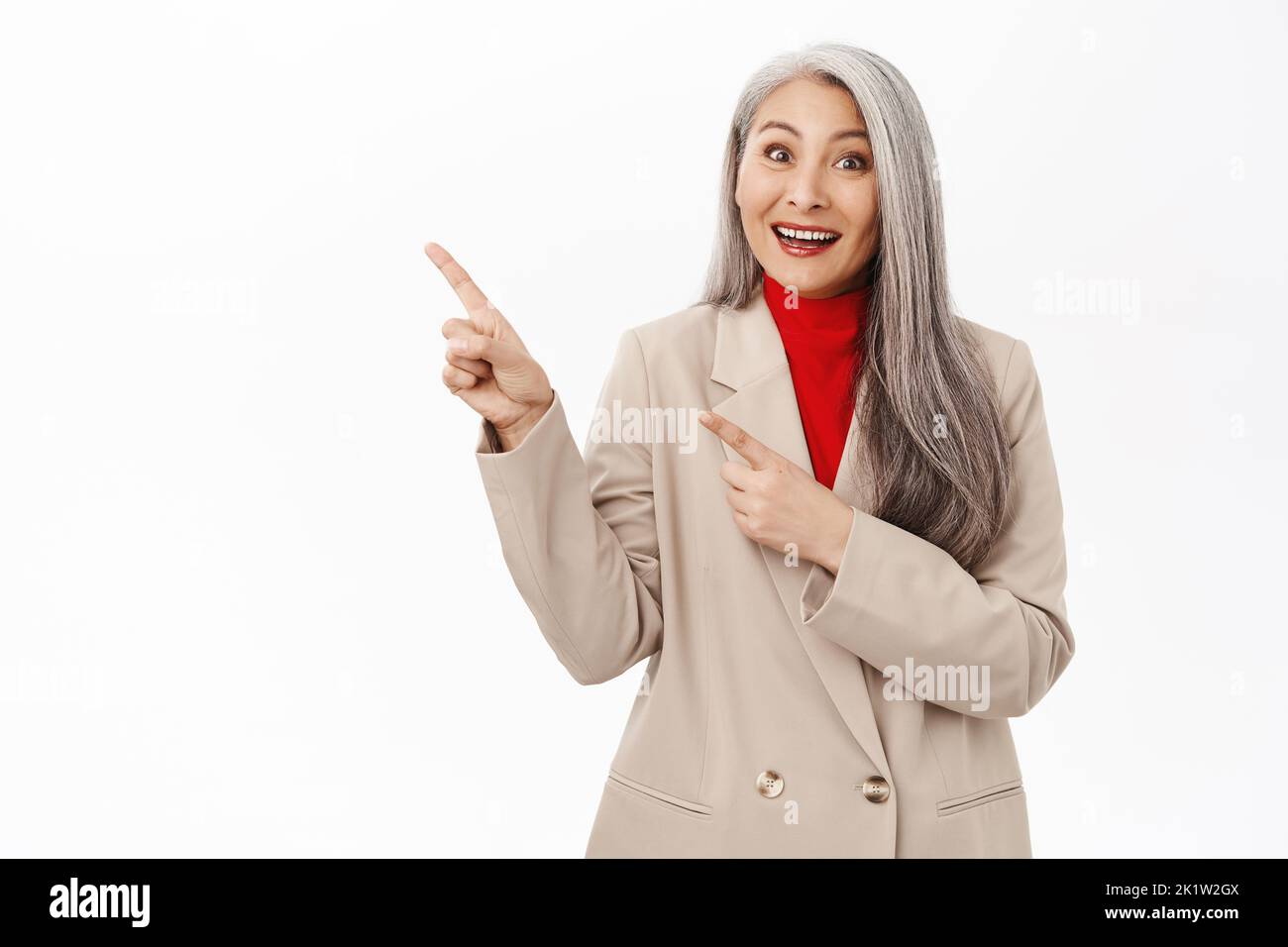 Beautiful senior asian businesswoman in suit pointing finger at upper left corner, smiling and looking amazed, showing advertisement, standing over wh Stock Photo