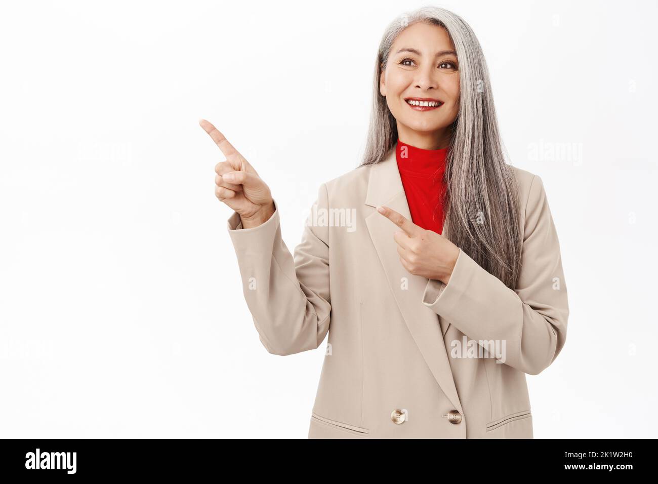 Image of smiling senior asian business lady, female entrepreneur pointing finger left at product banner and looking happy, standing over white backgro Stock Photo