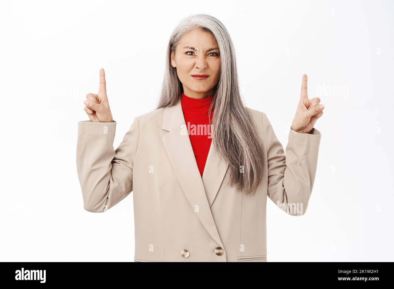 Image of displeased asian female entrepreneur, corporate woman pointing at product and grimacing upset, standing over white background. Stock Photo