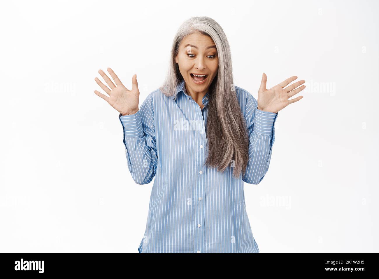 Excited asian senior woman, granny looking surprised down, feeling excited as winning, open gift, standing over white background. Stock Photo
