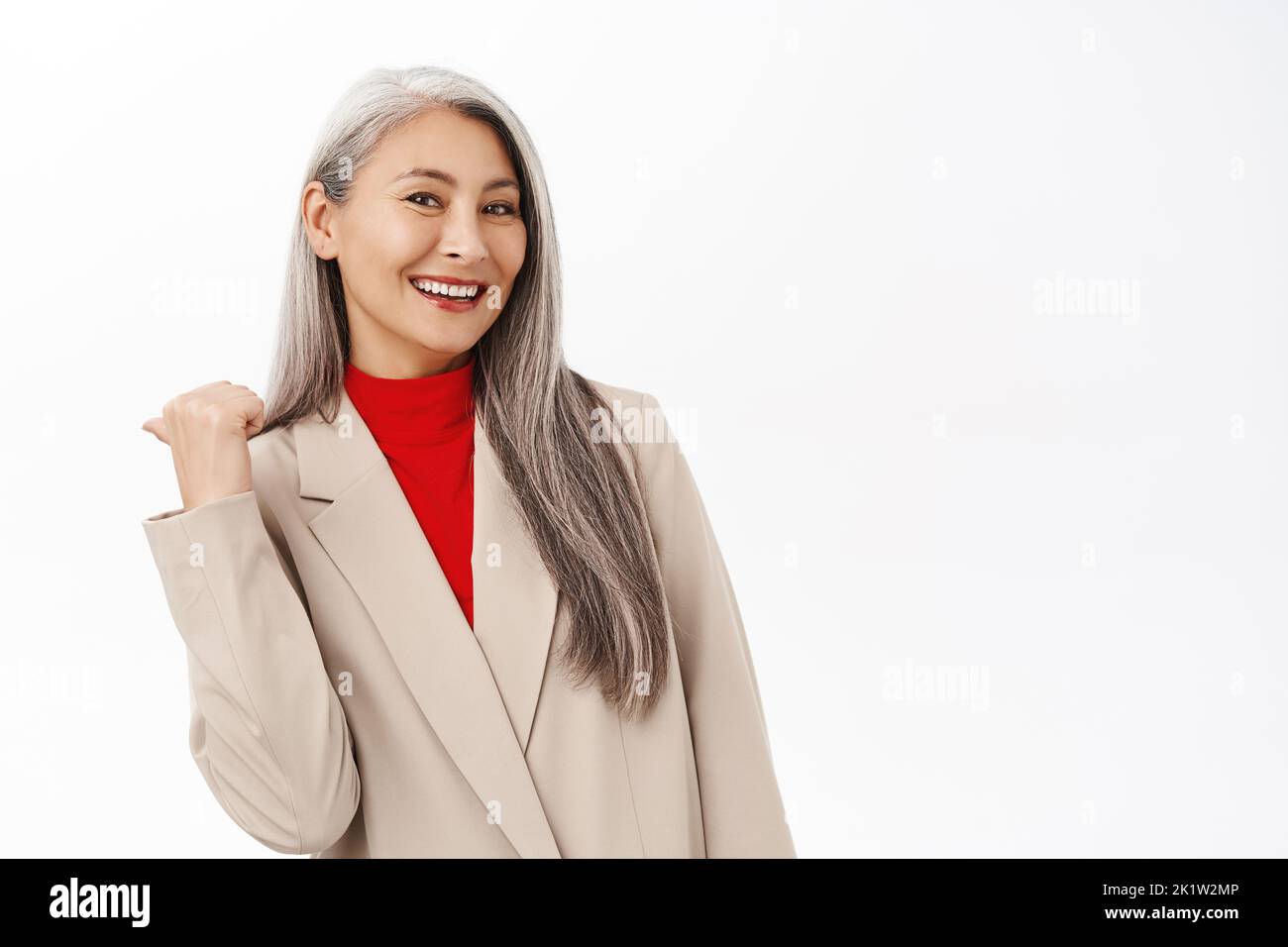 Portrait of asian businesswoman pointing behind her, showing smth on back, smiling and laughing, standing over white background. Corporate people conc Stock Photo