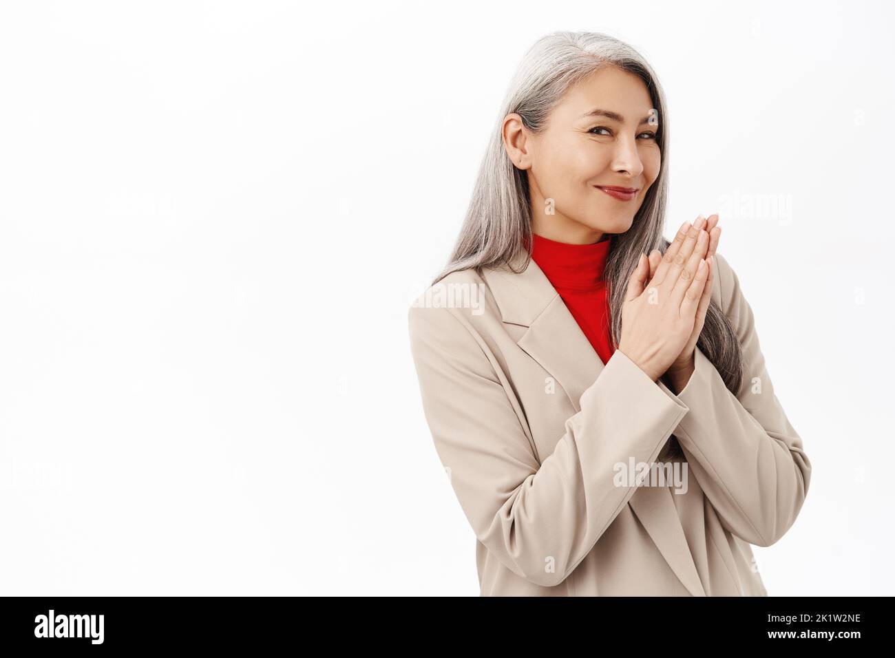 Portrait of smiling pleased asian senior woman, businesswoman rubbing hands with satisfied face expression, standing over white background. Stock Photo