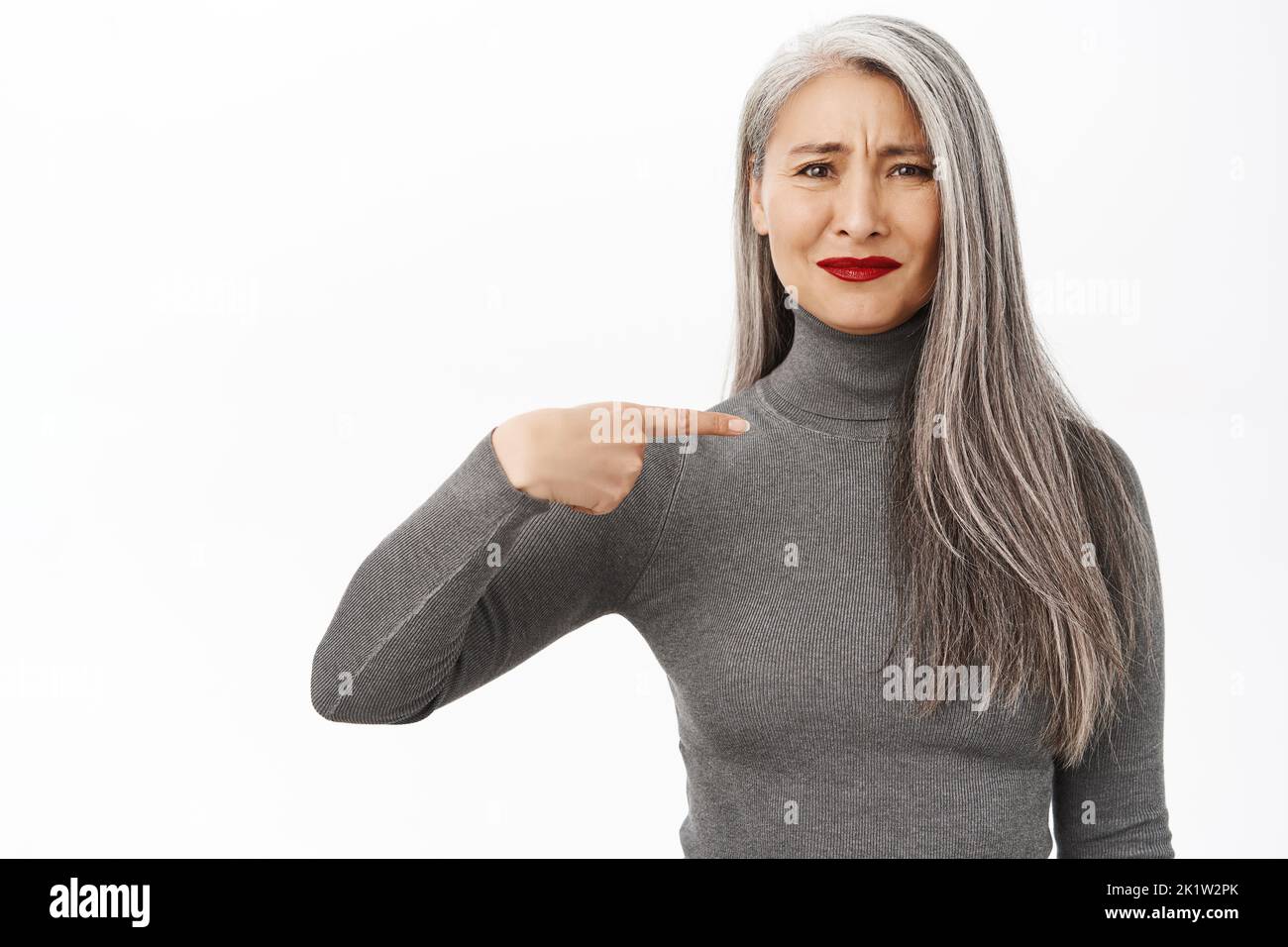Beautiful asian senior woman looking reluctant, pointing at herself with skeptical face, frowning and grimacing, standing over white background. Stock Photo