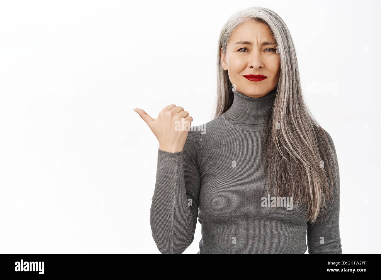 Portrait of smiling beautiful asian middle aged woman with red lipstick, looking at camera, pointing left at empty copy space for advertisement, stand Stock Photo