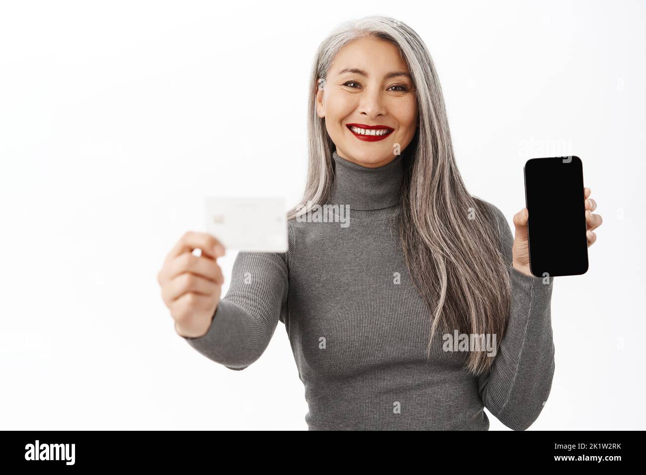 Bank and people concept. Happy asian lady showing mobile phone screen, credit card, demonstrates app interface on smartphone, standing over white back Stock Photo