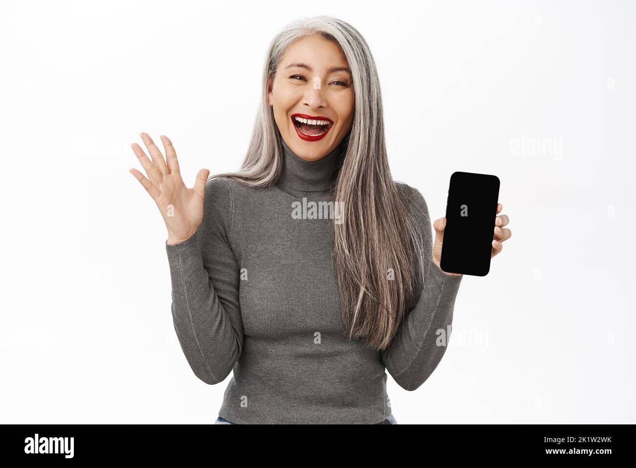 Portrait of excited asian senior woman showing mobile phone screen and celebrating, achieve goal on smartphone app, standing happy against white backg Stock Photo