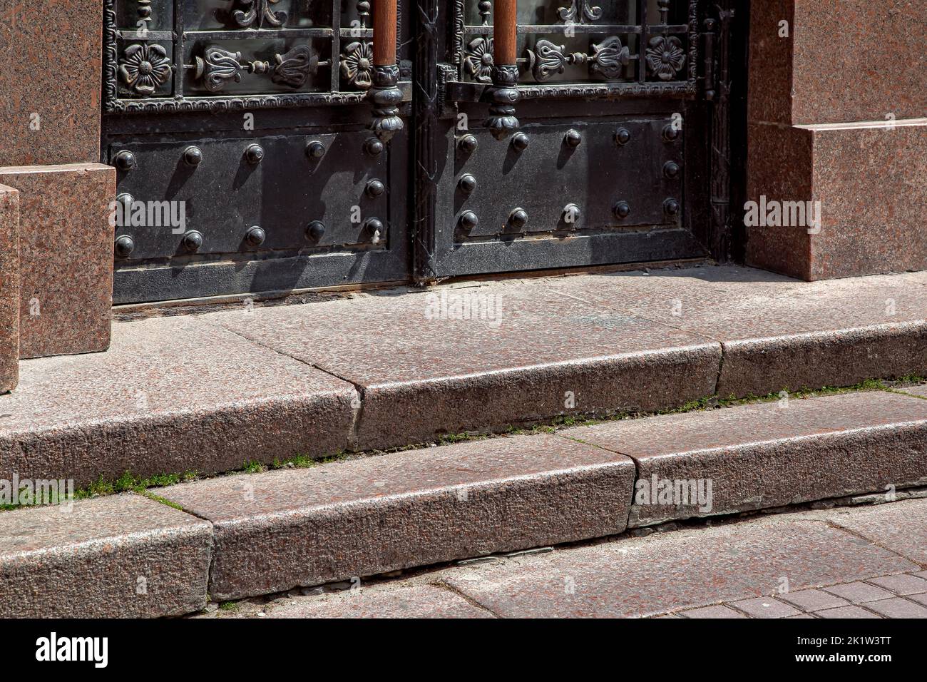 granite staircase with stone steps rise to the entrance black iron door with patterns and handles, building facade architecture lit by sun close-up, s Stock Photo