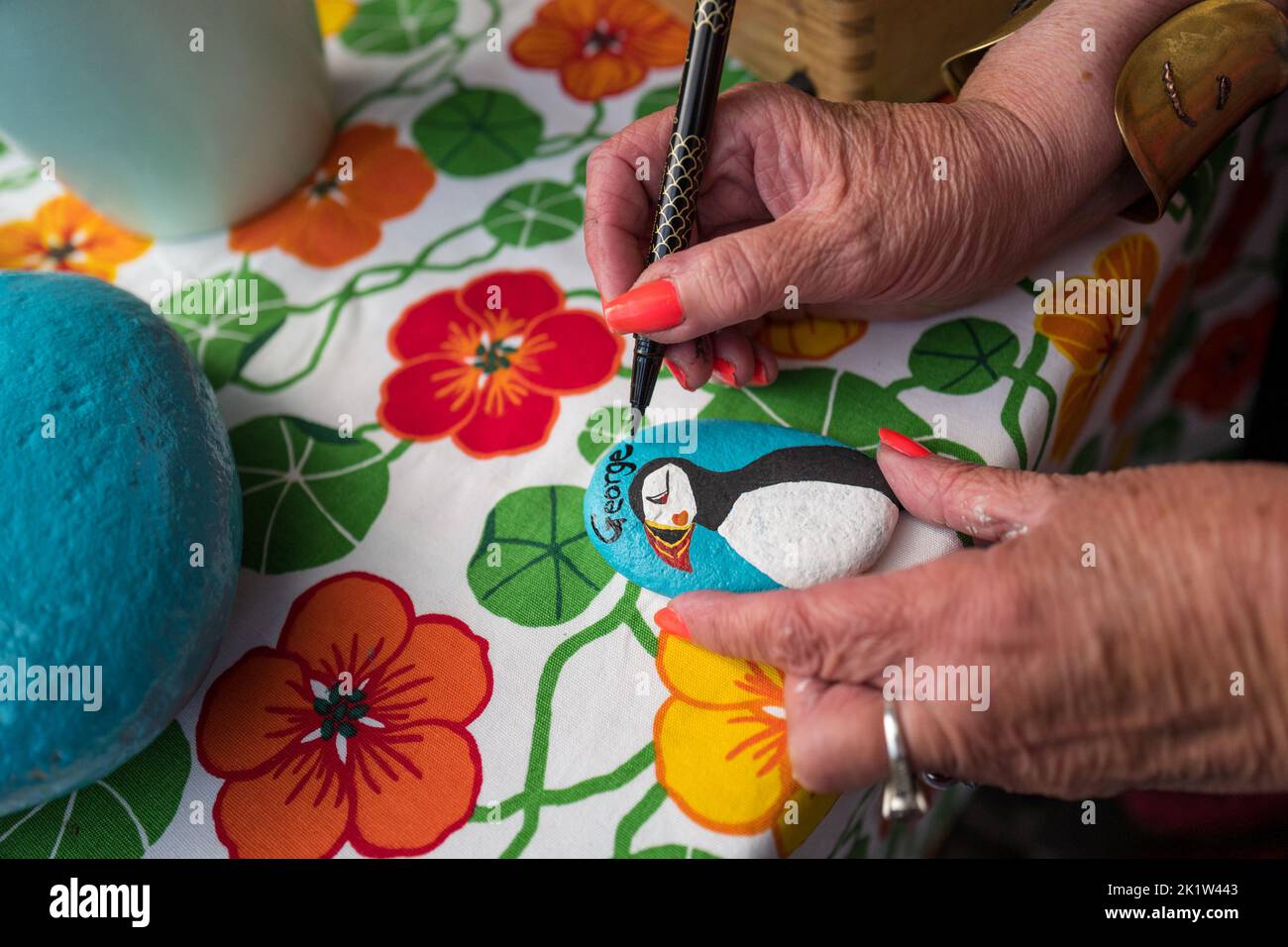 Female artist writing the name George with a black felt pen onto a painted stone with a puffin at a market stall. Stock Photo