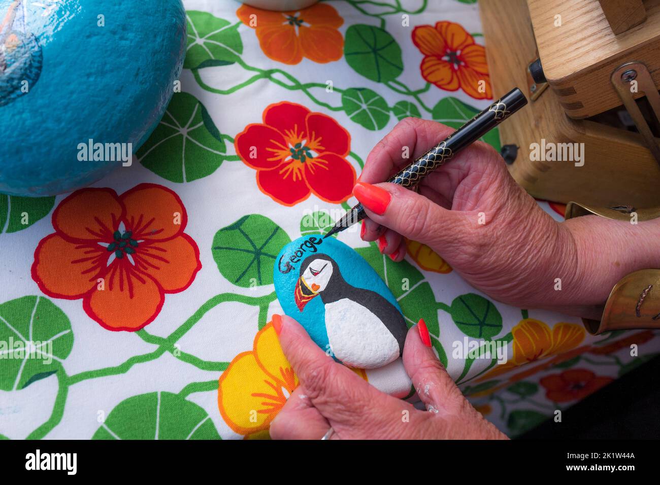 Female artist writing the name George with a black felt pen onto a painted stone with a puffin at a market stall. Stock Photo