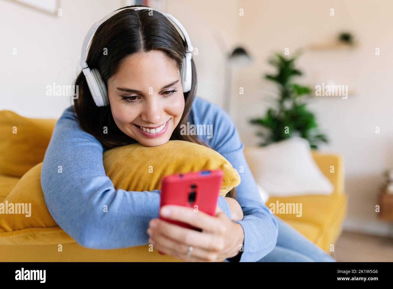 Smiling young woman with headphones using mobile phone lying on sofa at home Stock Photo
