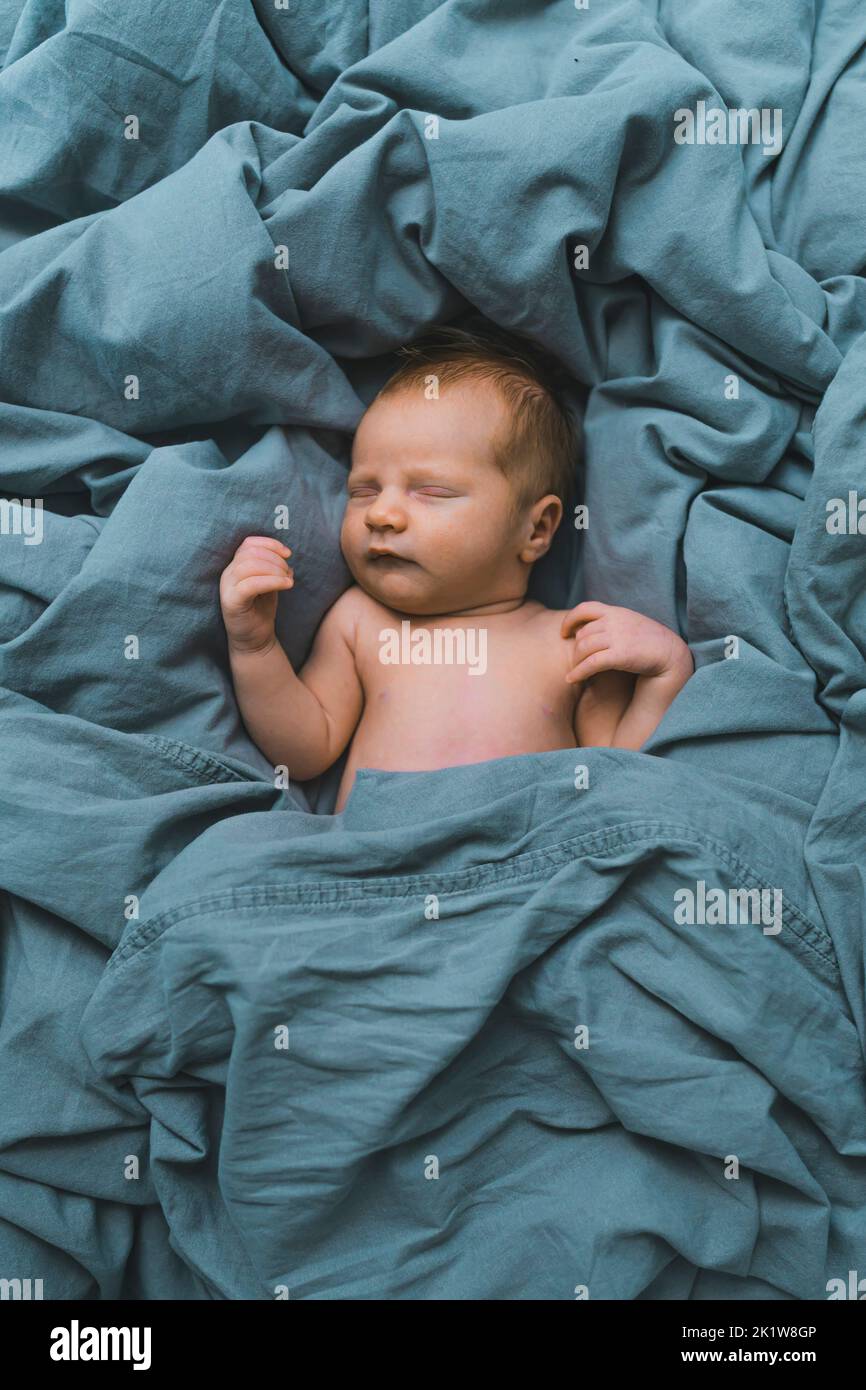 White newborn baby boy sleeping in blue bedsheets with arms up and