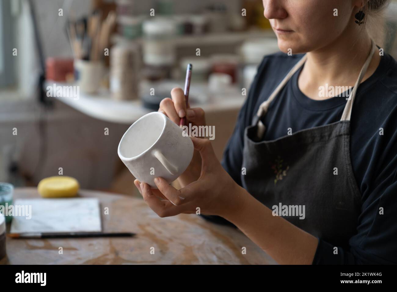 Craftswoman makes sketch of future pattern on white mug to decorate kitchen with clay dish Stock Photo