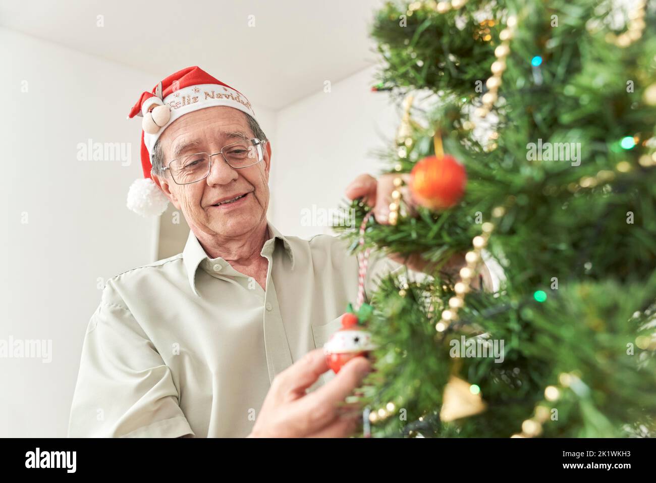 Happy senior hispanic man smiling while decorating a Christmas tree at home wearing a red Santa Claus beanie with a reindeer and the text Merry Christ Stock Photo