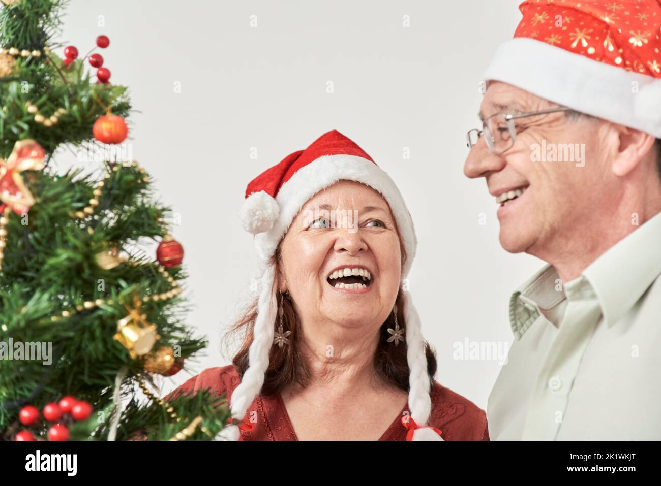 Happy hispanic senior couple smiling while decorating their Christmas tree, wearing red Santa Claus beanies. The joy of spending the holidays together Stock Photo