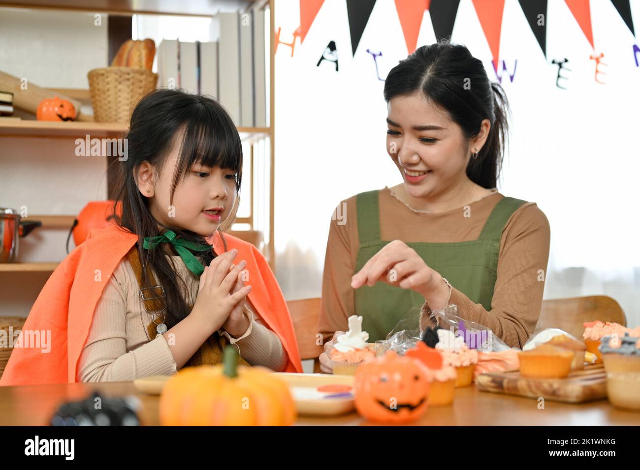 Happy and lovely young Asian girl in Halloween costume enjoys decorating cupcakes with her mom at home. Happy Halloween holiday concept Stock Photo