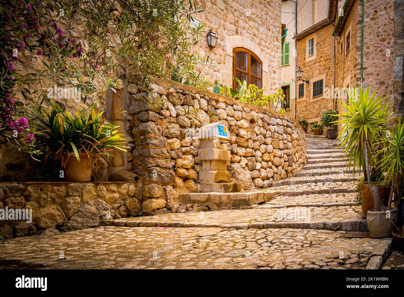 A narrow curved alley with cobbled steps leads through Mallorca mountain Village Biniaraix with mediterranean stone houses and a drinking water source Stock Photo