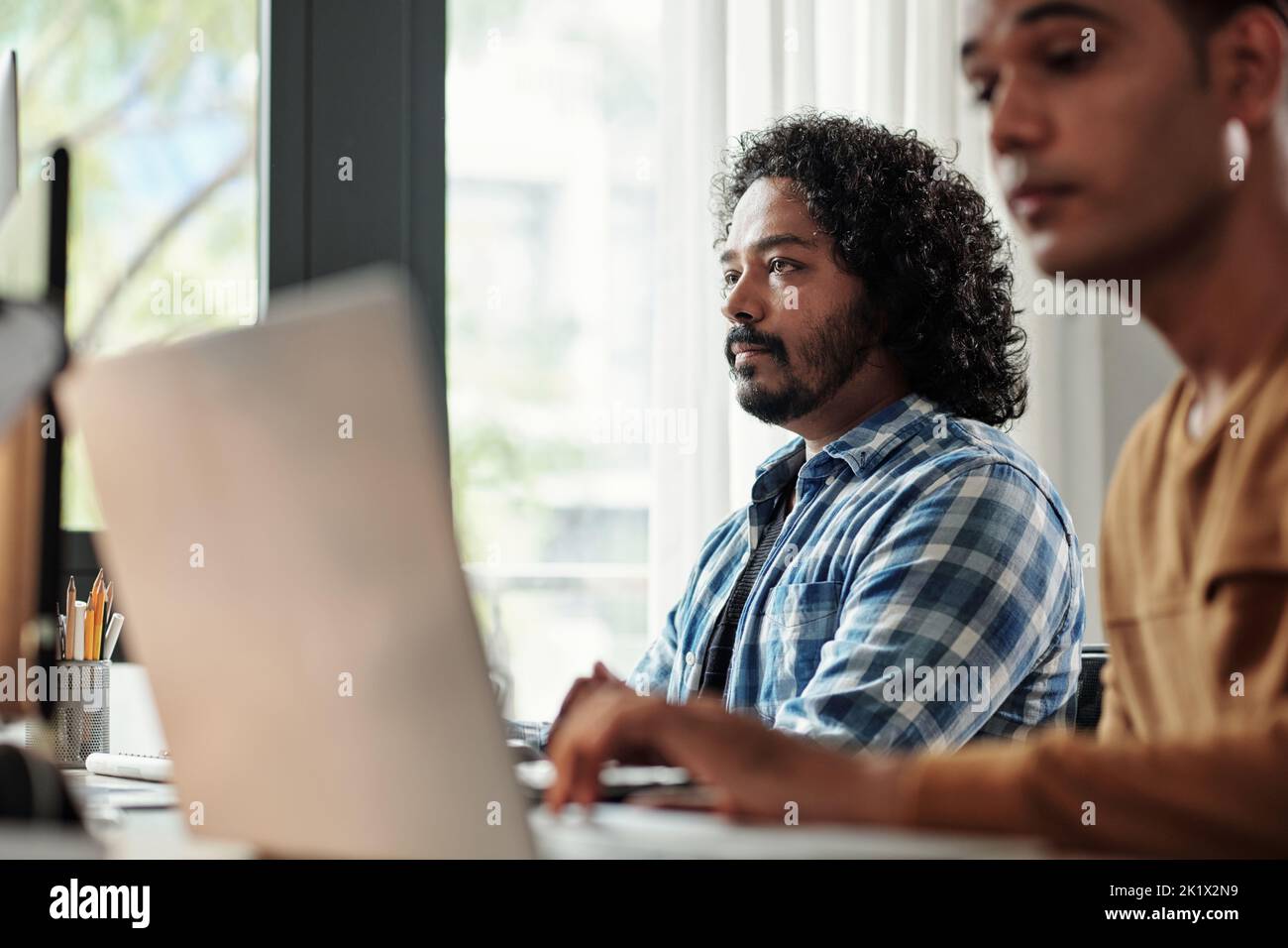 Pensive Indian programmer with curly hair looking at computer screen Stock Photo