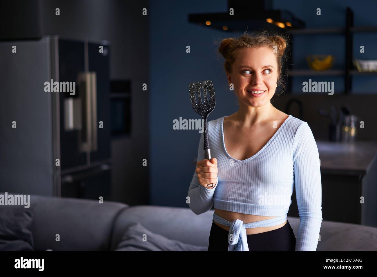 Young caucasian blonde woman holding black spatula smiling happy with teeth standing at home on dark modern stylish kitchen background. Young cheerful Stock Photo