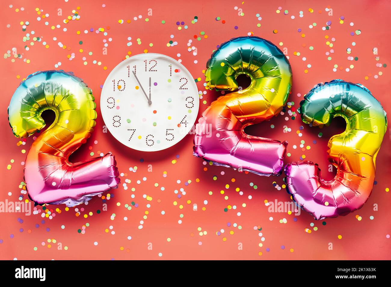 Colorful bright air balloon numbers and white watch at 12 o'clock on bright red confetti background. 2023 New Year concept celebration Stock Photo