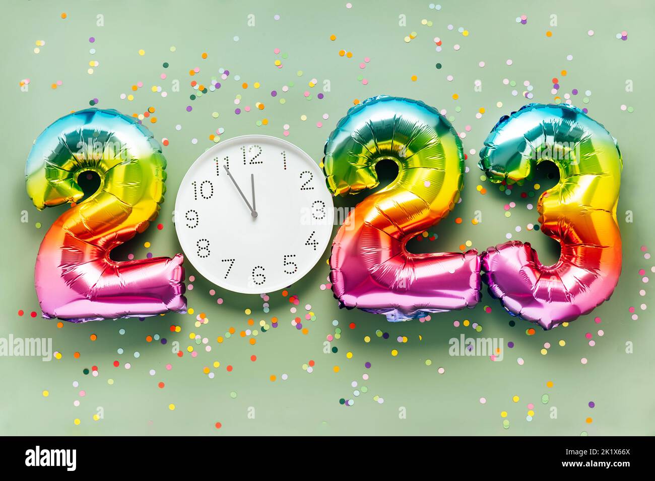 Colorful bright air balloon numbers and white watch at 12 o'clock on light green background with confetti. 2023 New Year concept celebration Stock Photo