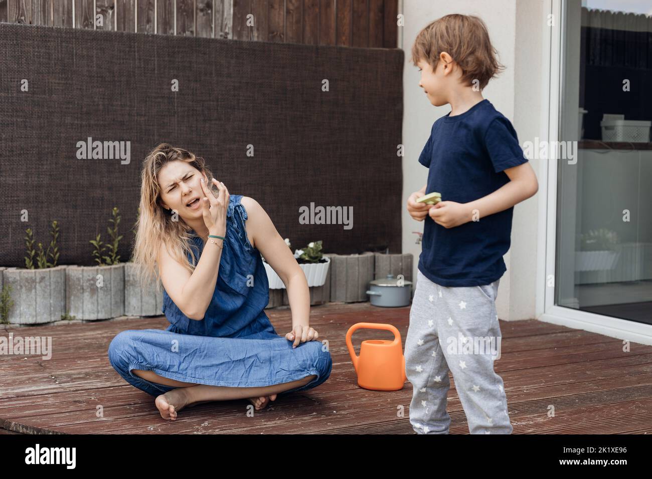 Blonde woman sit on bending legs and her son spend time on porch in countryside. Female itching her cheek, insect bite Stock Photo