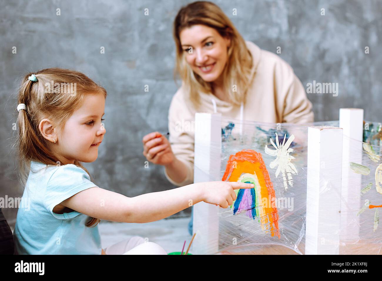Female teacher with little blonde girl painting rainbow on oilcloth. Fun learning in kid development childcare center Stock Photo