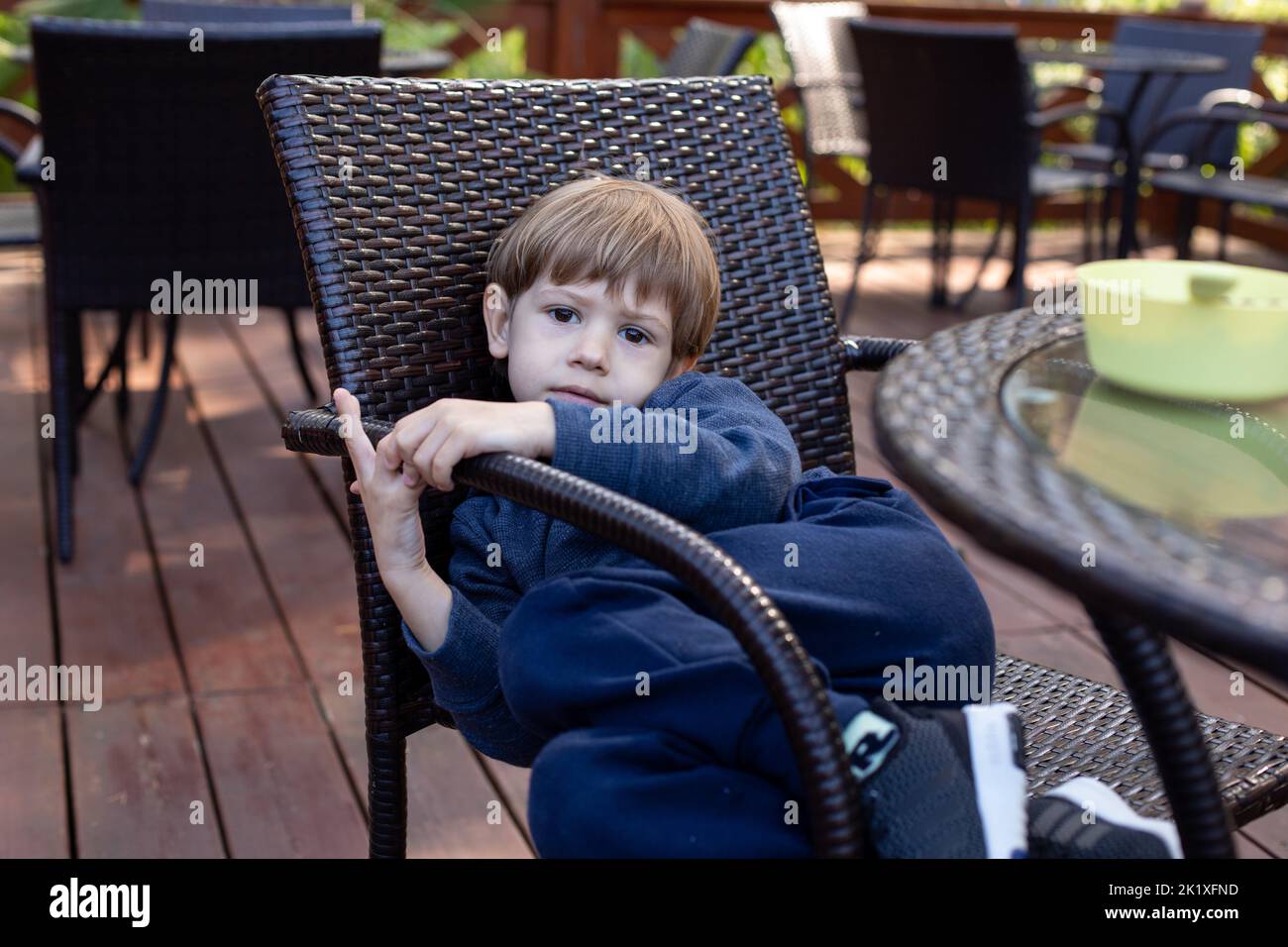 Unhappy little boy lie bending body on chair at table in cafe on summer terrace. School aged kid having dinner outdoor in public catering. Health Stock Photo