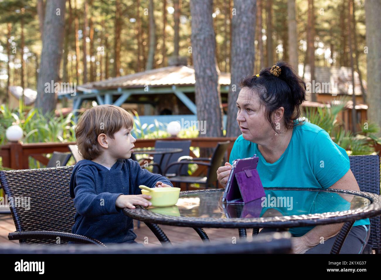 Little boy sit on chair at table on summer terrace and talk with mother. Kid of kindergarten age having dinner outdoor in forest. Communication with Stock Photo