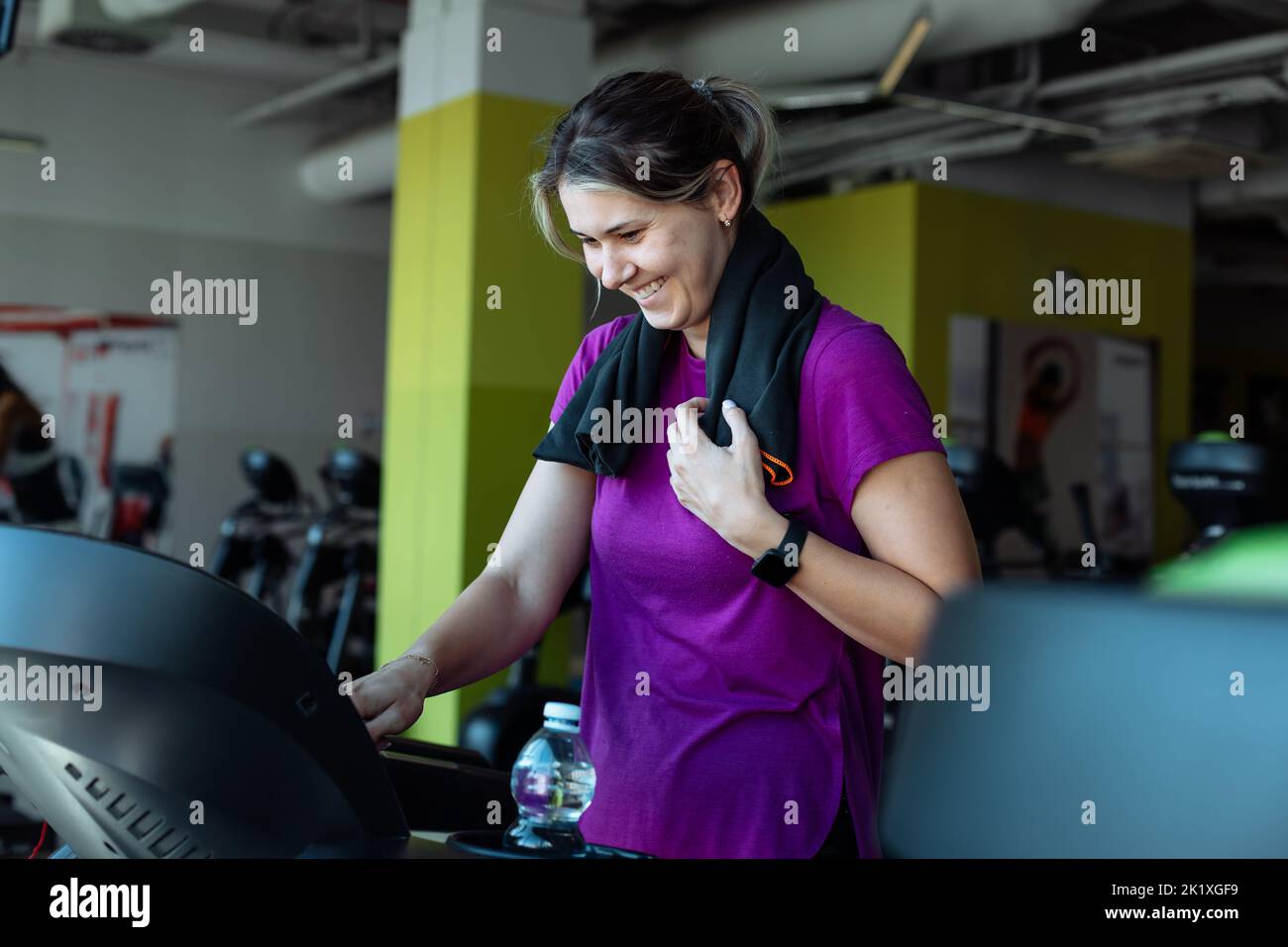 Happy overweight woman with smile run on treadmill at fitness gym with towel hanging around neck, side view. Young plus size girl do cardio workout on Stock Photo