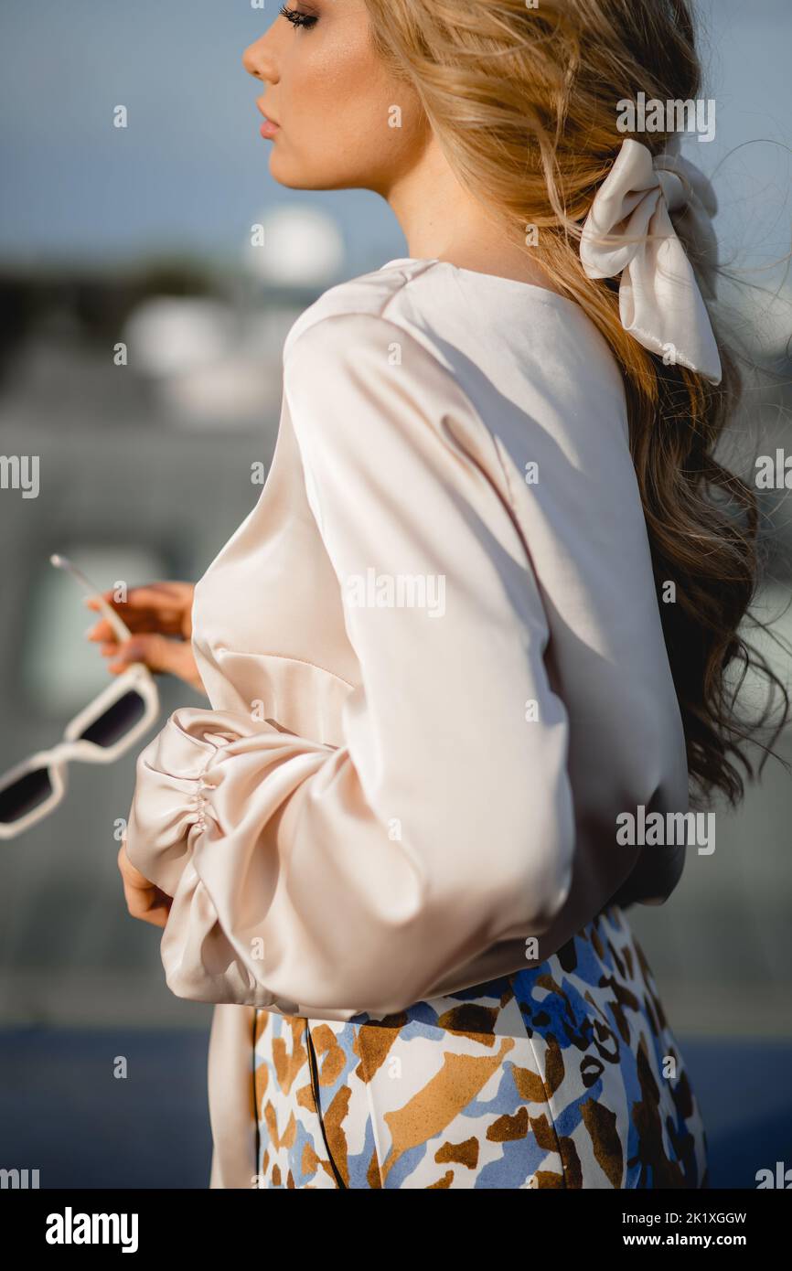 Gorgeous young woman in elegant silk blouse stand on roof terrace with sunglasses sideways closeup. Photo of parisian lady in stylish outfit on sky Stock Photo