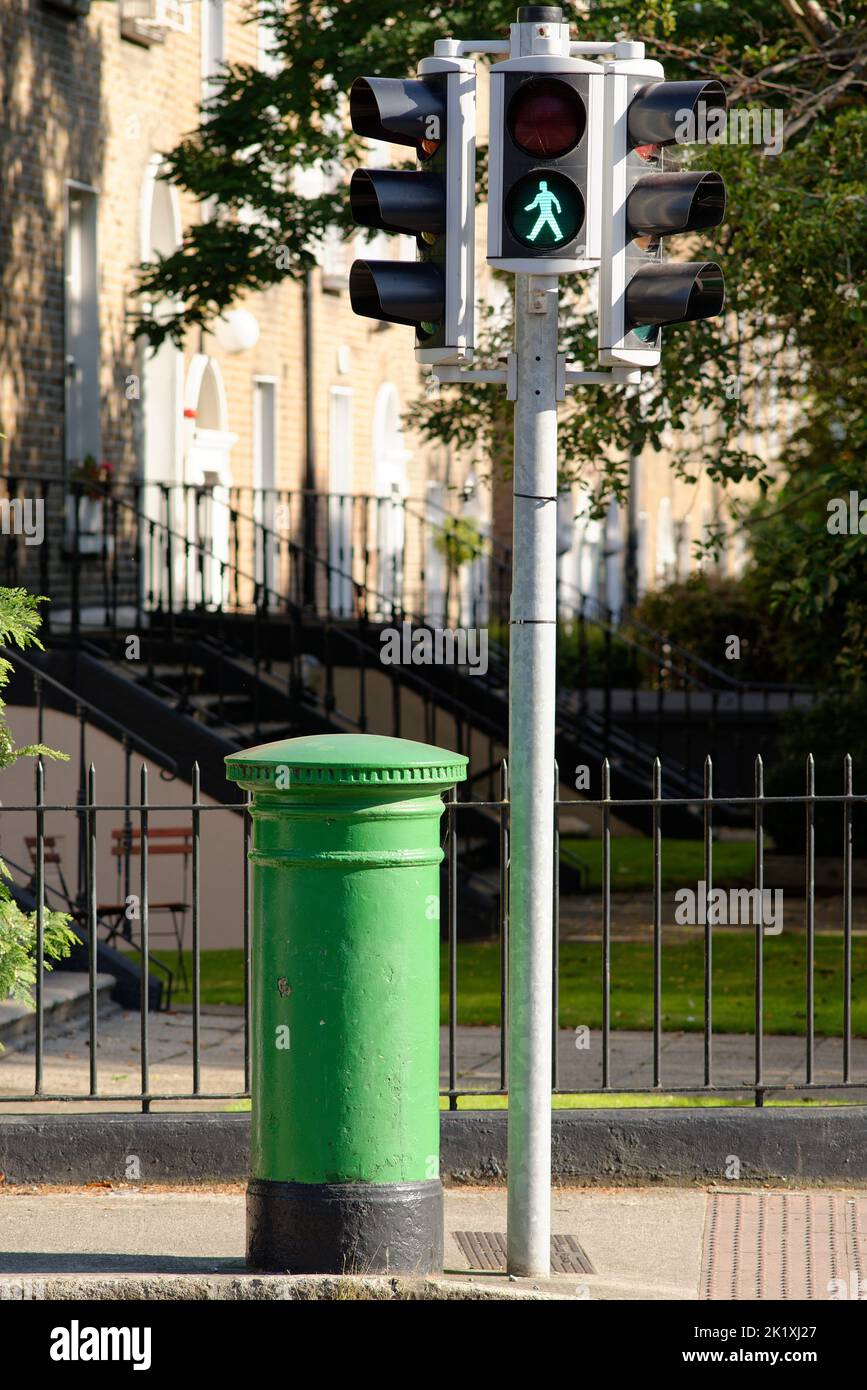 A traditional Irish letter box painted green. There is a green man 'Walk' signal on traffic lights above. A green post box for the postal service. Stock Photo