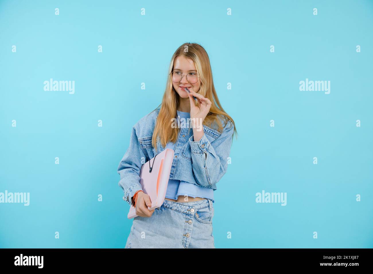 Confident, thoughtful, smiling blonde teenage girl in eyeglasses holding paper folder and biting pen. Back to school Stock Photo