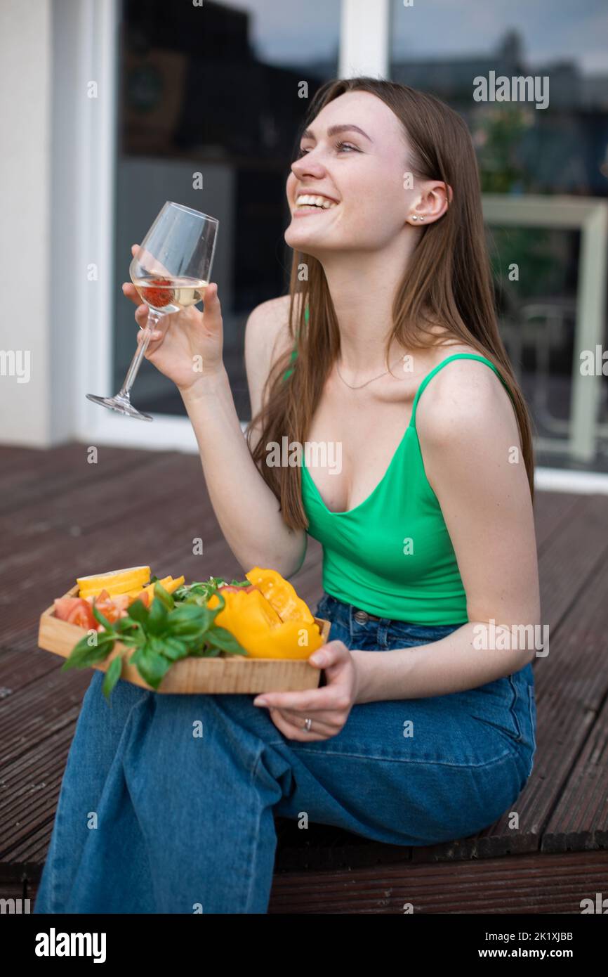 Vertical laughing brunette woman in green top and jeans drink wine and eating tasty food on plate, sit on wooden floor Stock Photo