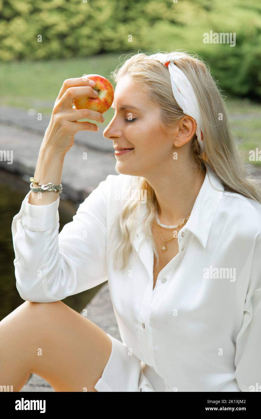 Vertical smiling, satisfied blonde stylish beautiful woman in white dress hold apple with close eyes in park near lake Stock Photo