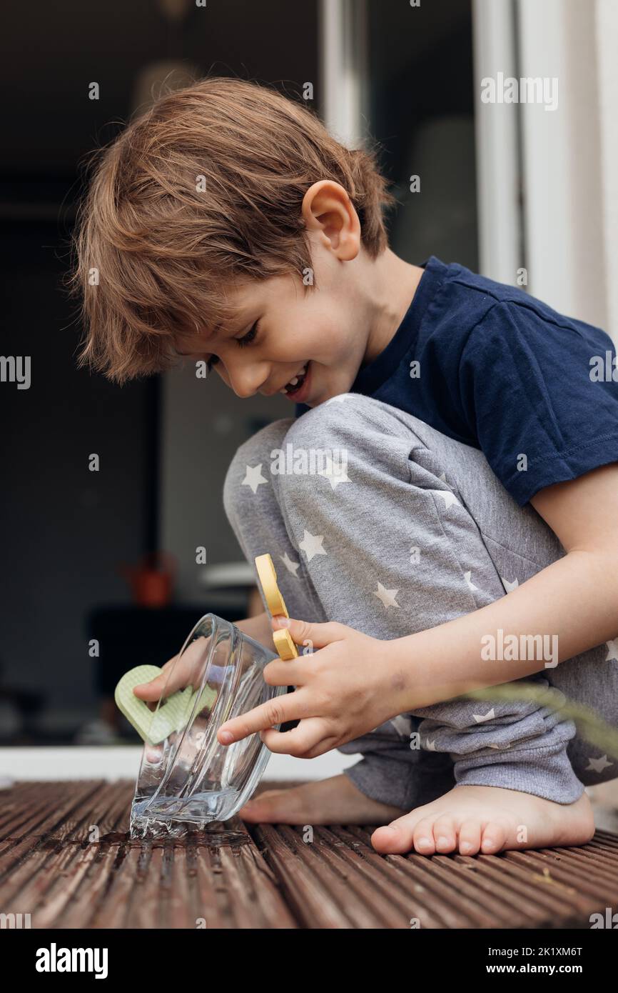 Vertical photo of little barefoot fair haired smiling boy washing and pouring clean water in animal bowl for his dog  Stock Photo