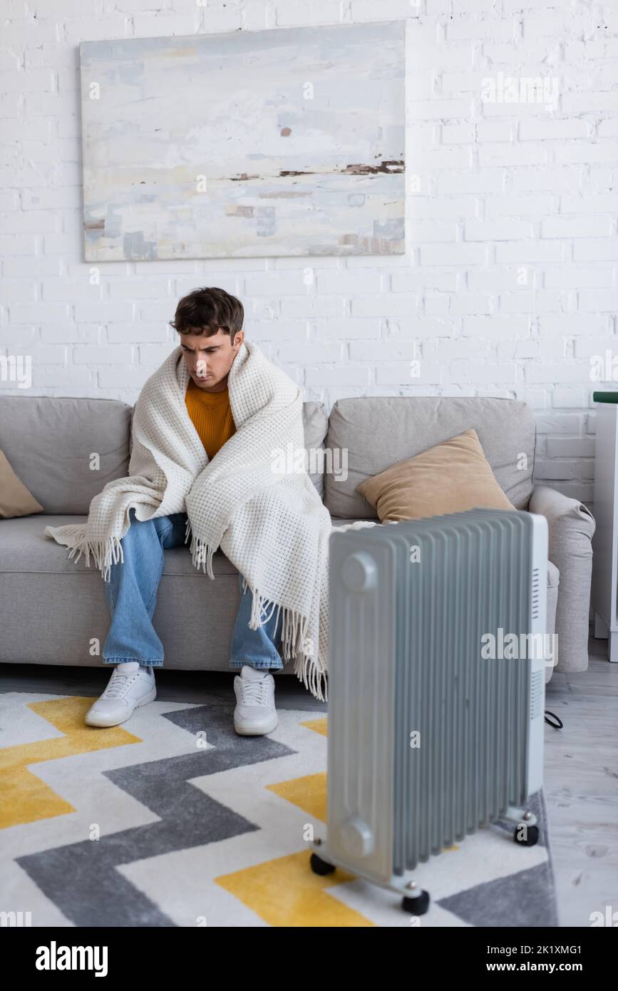 full length of young man covered in blanket sitting on sofa near radiator heater in winter,stock image Stock Photo