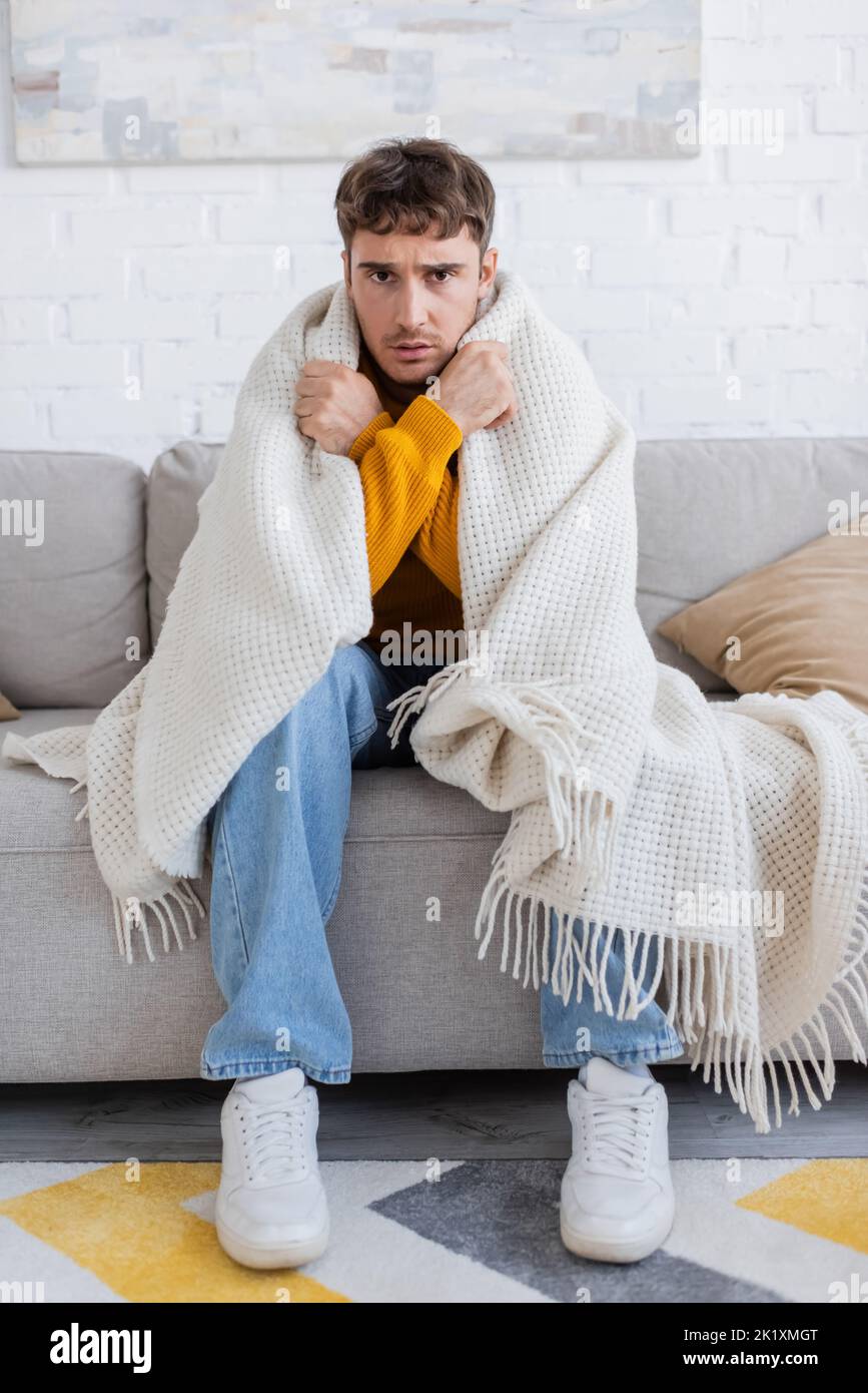 frustrated young man covered in blanket sitting on sofa while getting warm in living room,stock image Stock Photo