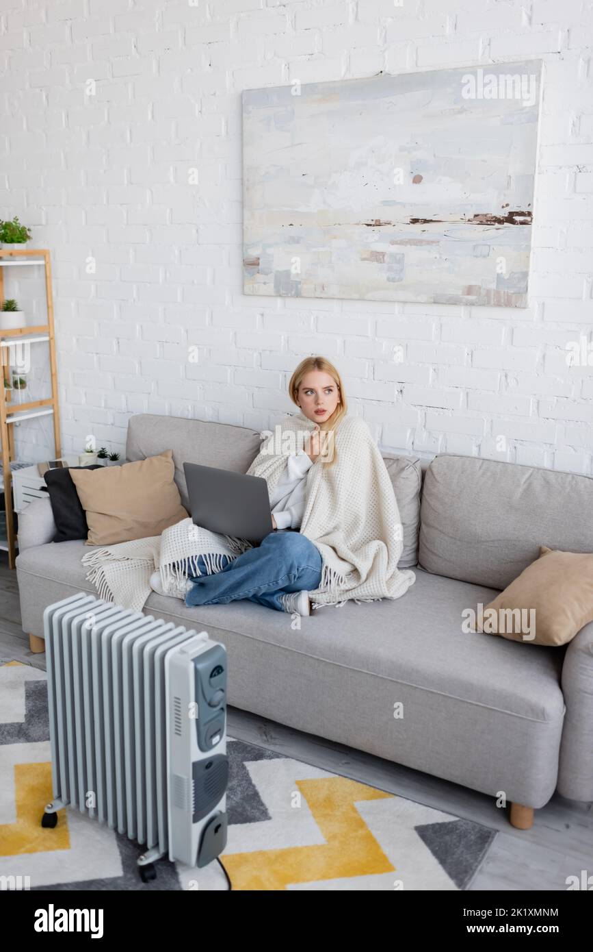 young blonde woman covered in white blanket using laptop while sitting ...