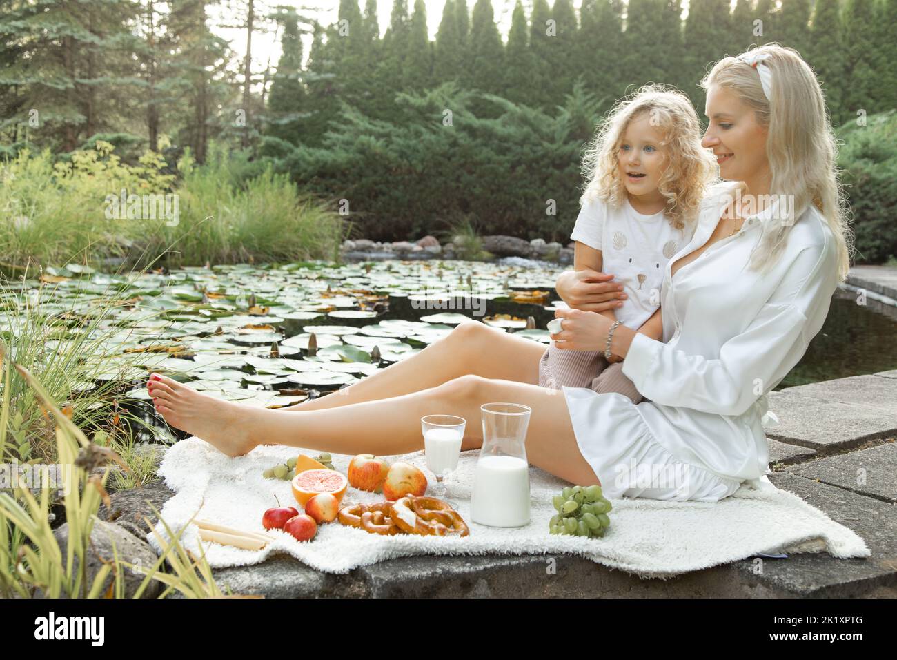 Joyful, smiling woman hold little blonde girl, have lunch on picnic in park near lake outdoors. Healthy ecological food Stock Photo