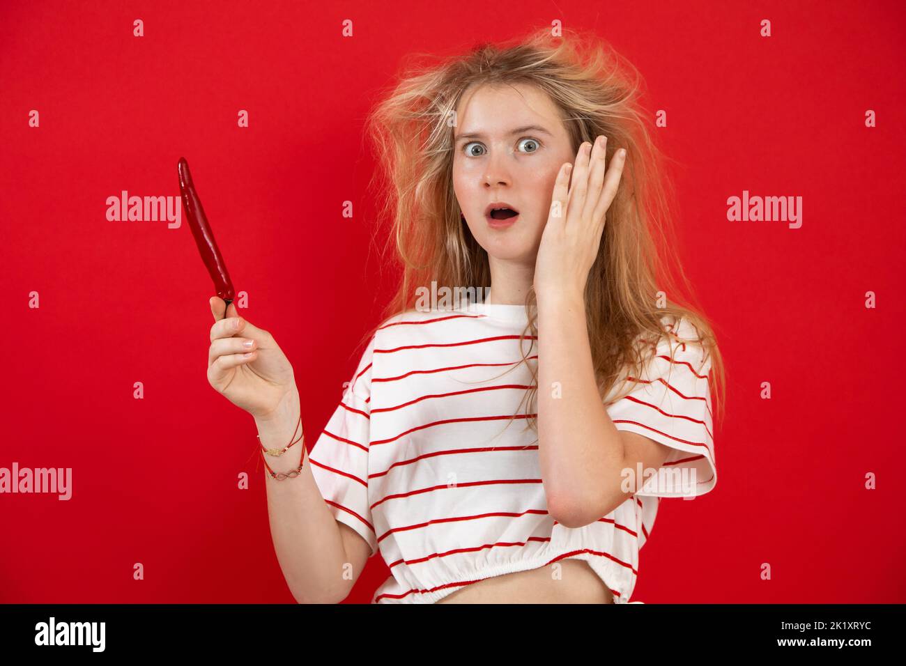 Expressive, wondering, surprised blonde teenage girl look at camera and hold red chili pepper in hand on red background Stock Photo
