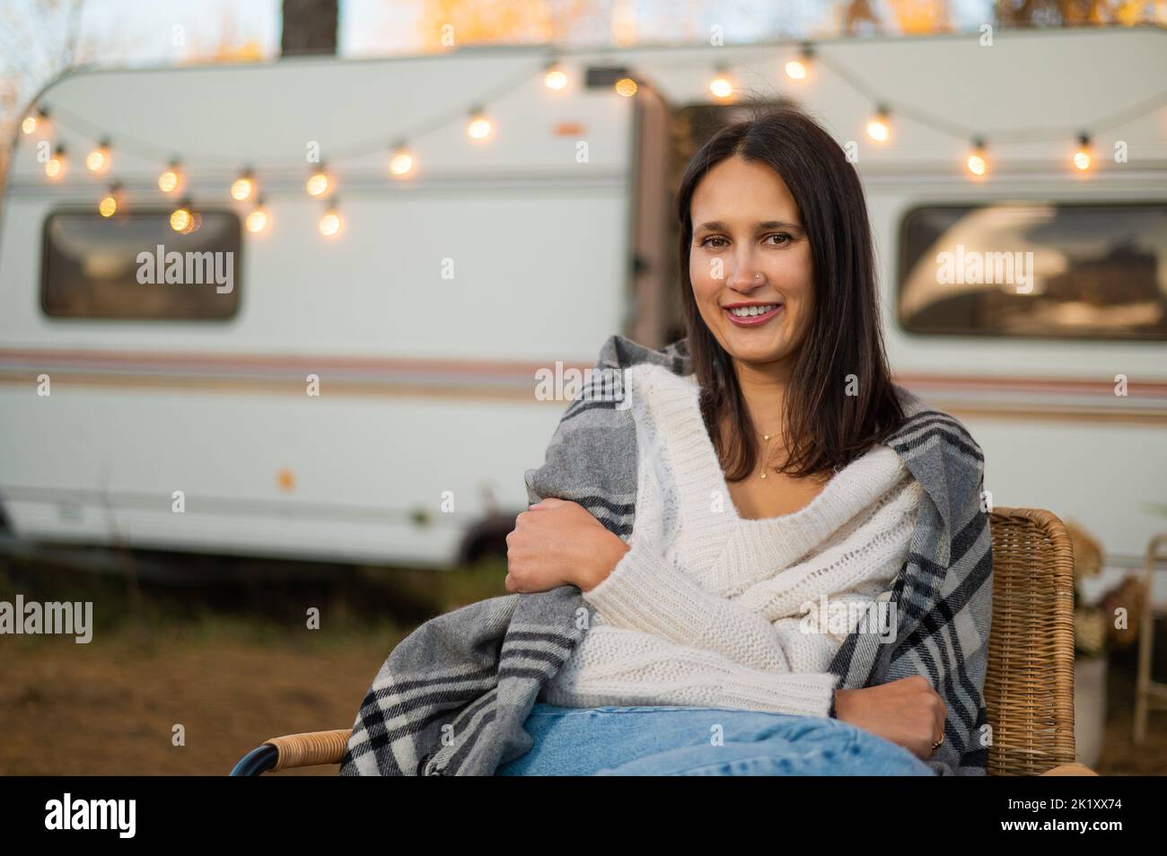 Caucasian woman sits in a wicker chair wrapped in a blanket in the yard near the trailer in autumn.  Stock Photo